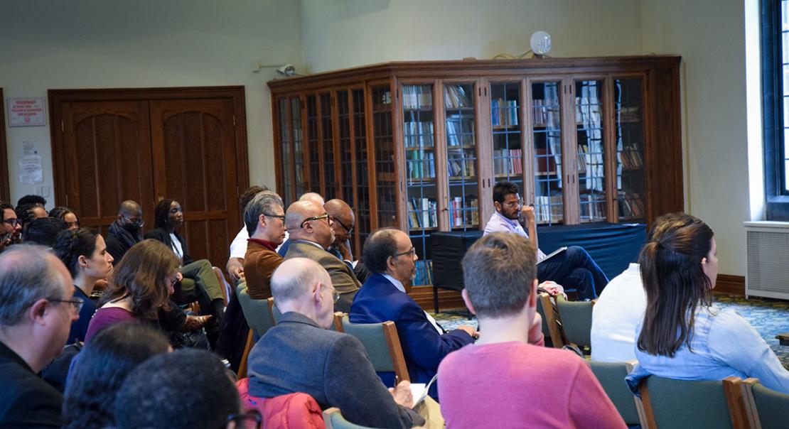 People are seated in a semi-circle in green chairs, listening and writing notes.